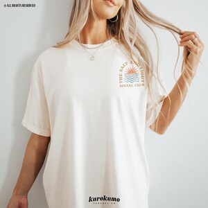 May include: A white t-shirt with a graphic of a sun setting over the ocean with the text "The Salt and Light Social Club".