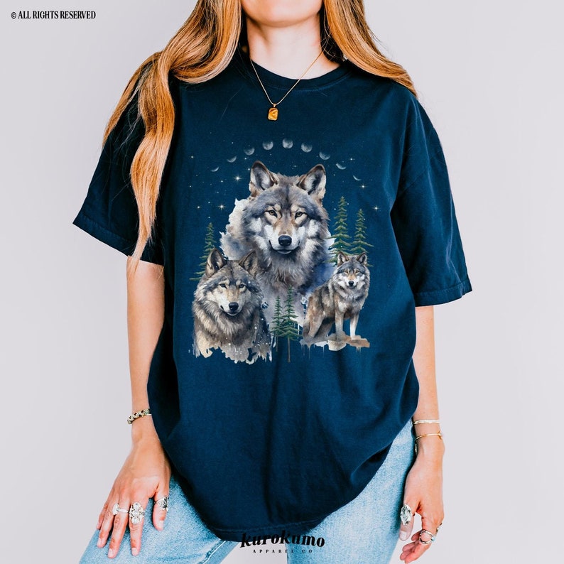 May include: A navy blue t-shirt with a graphic print of three wolves in a forest setting. The wolves are surrounded by trees and a crescent moon. The graphic is in shades of gray and white.