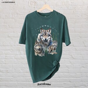 May include: A dark green t-shirt with a watercolor design of three wolves in a forest setting. The wolves are gray and white, and the forest is a mix of green and blue.
