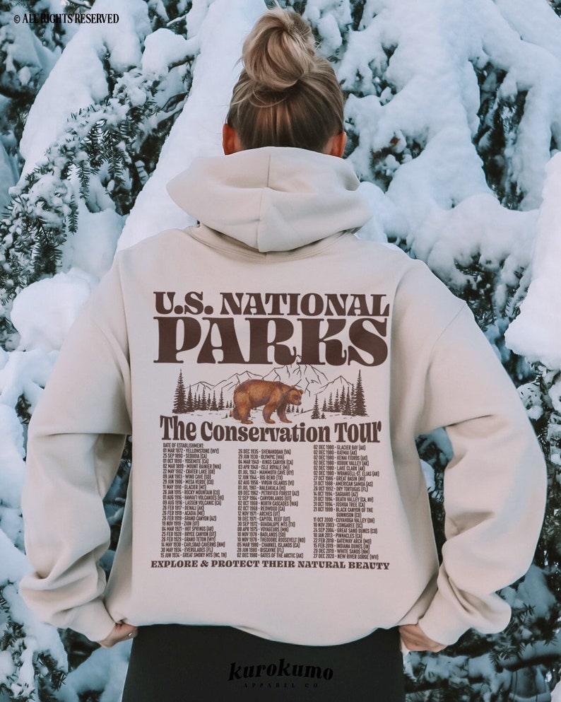 Pu&ograve; includere: Una felpa con cappuccio beige con la scritta "U.S. NATIONAL PARKS" e "The Conservation Tour" stampata sul retro. Il design include un orso e un elenco di date. La felpa viene indossata all'aperto in un ambiente innevato.