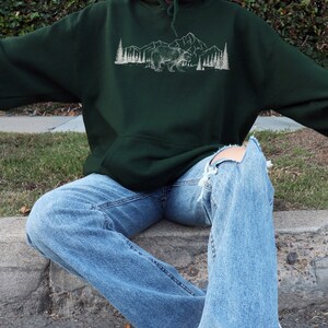 Pu&ograve; includere: Felpa con cappuccio verde scuro con grafica bianca di un orso, montagne e alberi. La felpa ha una tasca frontale e un cappuccio con coulisse. La persona indossa jeans blu chiaro con ginocchia strappate. Il marchio "kurokumo apparel co" &egrave; in basso.