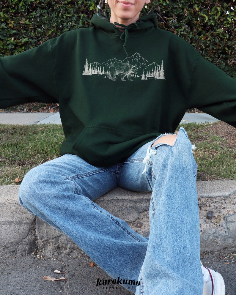 Pu&ograve; includere: Felpa con cappuccio verde scuro con grafica bianca di un orso, montagne e alberi. La felpa ha una tasca frontale e un cappuccio con coulisse. La persona indossa jeans blu chiaro con ginocchia strappate. Il marchio "kurokumo apparel co" &egrave; in basso.