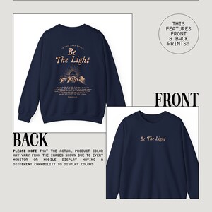 May include: Navy blue sweatshirt with a front and back print. The front print says "Be The Light" in a light peach color. The back print features a mountain scene with the text "In this dark world, Be The Light" and a bible verse.