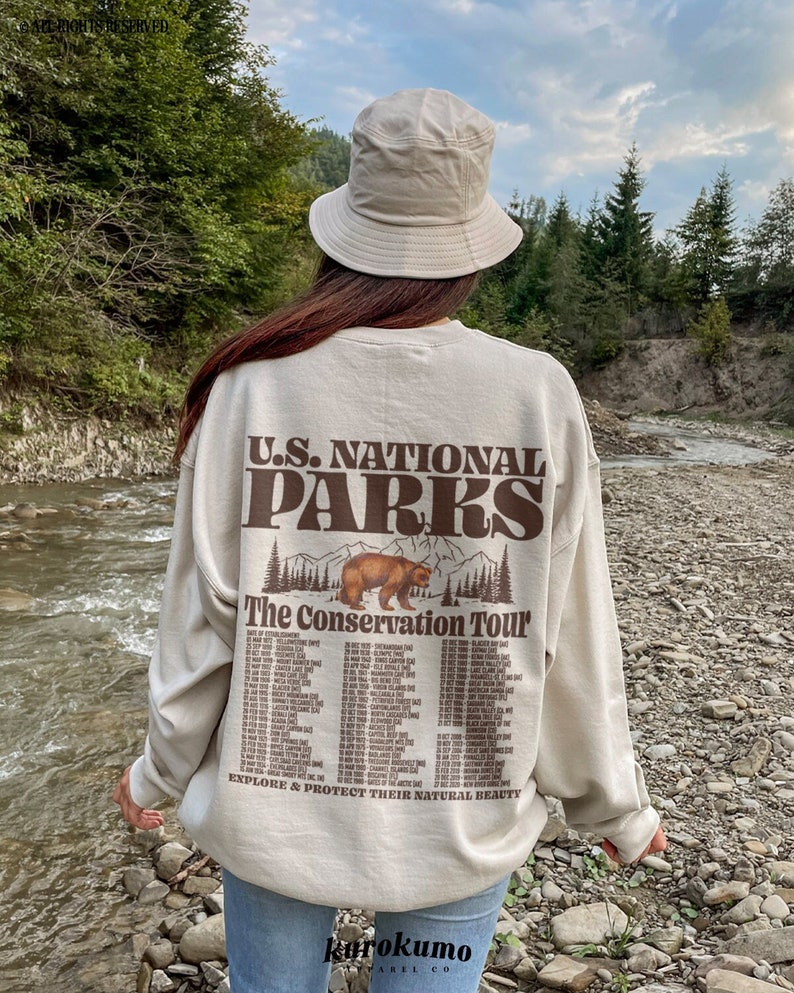 May include: A beige sweatshirt with the text "U.S. NATIONAL PARKS" and "The Conservation Tour" in brown. A bear graphic is above the text. The wearer is also wearing a beige bucket hat. The background is a river and trees.