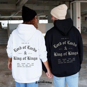 May include: Two people wearing black and white hooded sweatshirts with the text "For He Is Lord of Lords & King of Kings" printed on the back. The text is in a gothic font.