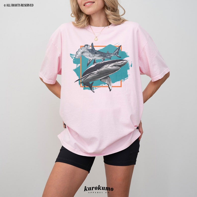 May include: Pink t-shirt with a graphic print of two hammerhead sharks and a shark in a teal blue and orange frame.