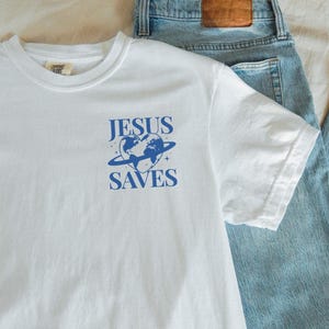 K&ouml;nnte beinhalten: Wei&szlig;es T-Shirt mit einem blauen Grafikdesign, das eine herzf&ouml;rmige Erde mit einem Ring darum und den Text "JESUS SAVES" zeigt. Das Shirt tr&auml;gt auch den Text "kurokumo APPAREL CO" unten.