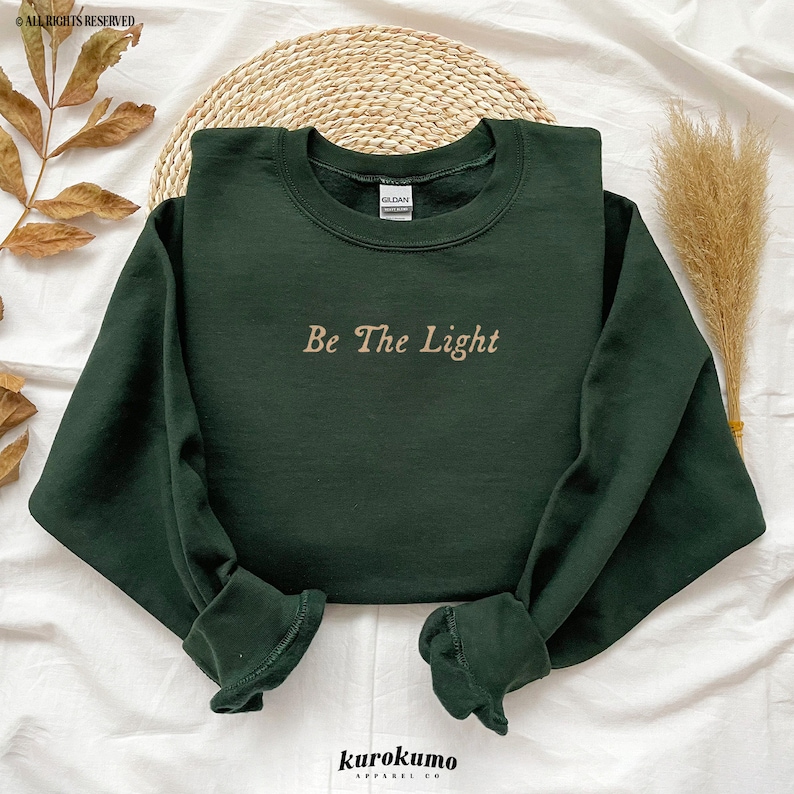 May include: A dark green sweatshirt with the text "Be The Light" in a light brown color.