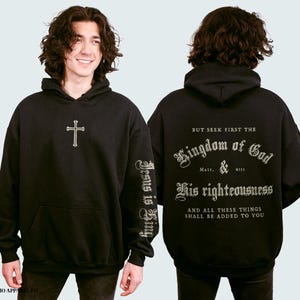 Op de afbeelding: Zwart hoodie met een kruis op de voorkant en de tekst "Jesus is King" op de linkermouw. De achterkant van de hoodie heeft de tekst "But seek first the Kingdom of God & His righteousness and all these things shall be added to you" in een decoratief lettertype.