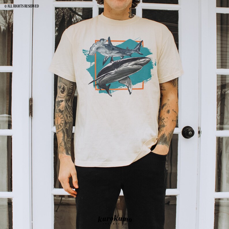 May include: A beige t-shirt with a graphic print of two sharks. The sharks are in a blue and orange abstract design. The shirt has short sleeves and a crew neck.