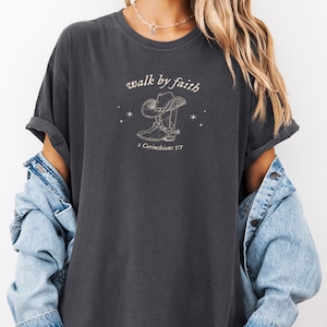 Pu&ograve; includere: Una t-shirt color antracite con una grafica di uno stivale da cowboy e un cappello con il testo "walk by faith" e "2 Corinthians 5:7".