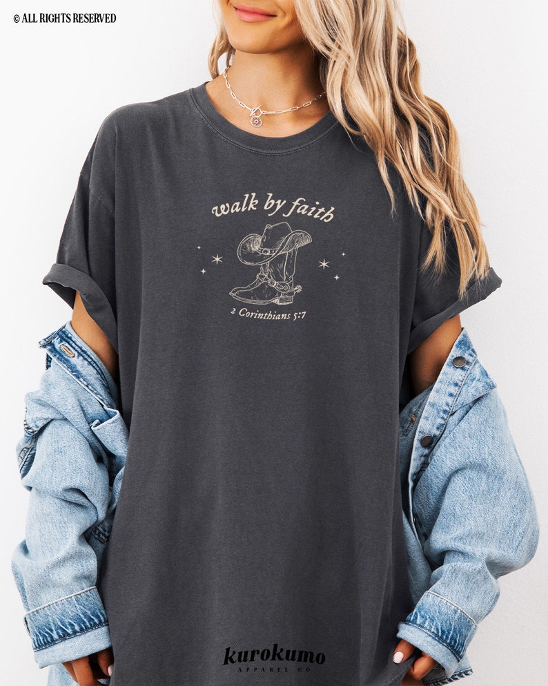 Pu&ograve; includere: Una t-shirt color antracite con una grafica di uno stivale da cowboy e un cappello con il testo "walk by faith" e "2 Corinthians 5:7".