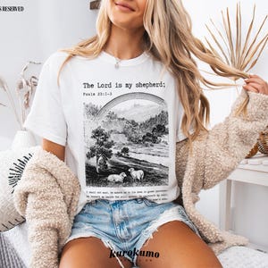 Puede incluir: Camiseta blanca con una ilustraci&oacute;n en blanco y negro de un paisaje y ovejas. El texto "The Lord is my shepherd; Psalm 23:1-3" est&aacute; impreso en la camiseta.