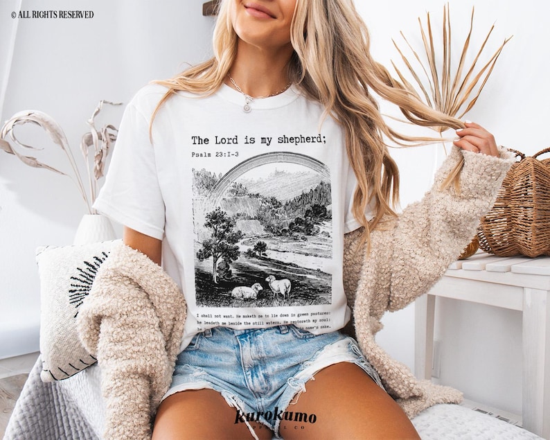 Puede incluir: Camiseta blanca con una ilustraci&oacute;n en blanco y negro de un paisaje y ovejas. El texto "The Lord is my shepherd; Psalm 23:1-3" est&aacute; impreso en la camiseta.