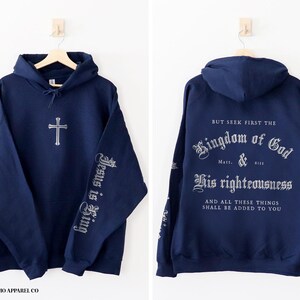 Op de afbeelding: Een marineblauw hoodie met de tekst "BUT SEEK FIRST THE Kingdom of God Matt. 6:33 His righteousness AND ALL THESE THINGS SHALL BE ADDED TO YOU" op de rug en "Jesus is King" op de linkermouw.