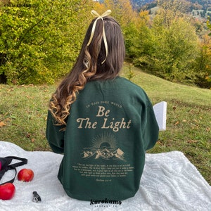 May include: A dark green sweatshirt with the text "In this dark world, Be the Light" printed on the back. The text is in a light brown color and is surrounded by a graphic of mountains and a sun. The sweatshirt is being worn by a person sitting on a grassy hill.