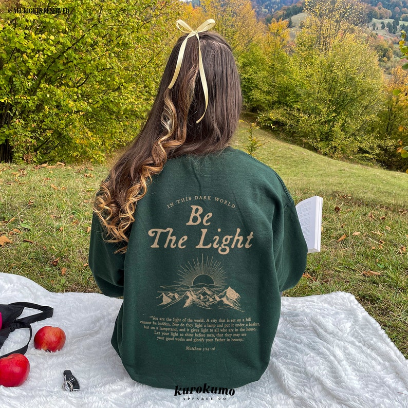 May include: A dark green sweatshirt with the text "In this dark world, Be the Light" printed on the back. The text is in a light brown color and is surrounded by a graphic of mountains and a sun. The sweatshirt is being worn by a person sitting on a grassy hill.