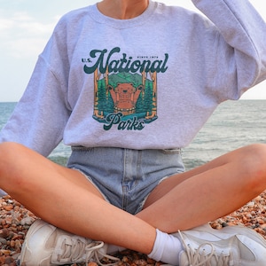 Może przedstawiać: Jasnoszara bluza z nadrukiem przedstawiającym niedźwiedzia w leśnej scenerii oraz napisem "U.S. National Parks Since 1872". Bluza ma okrągły dekolt.