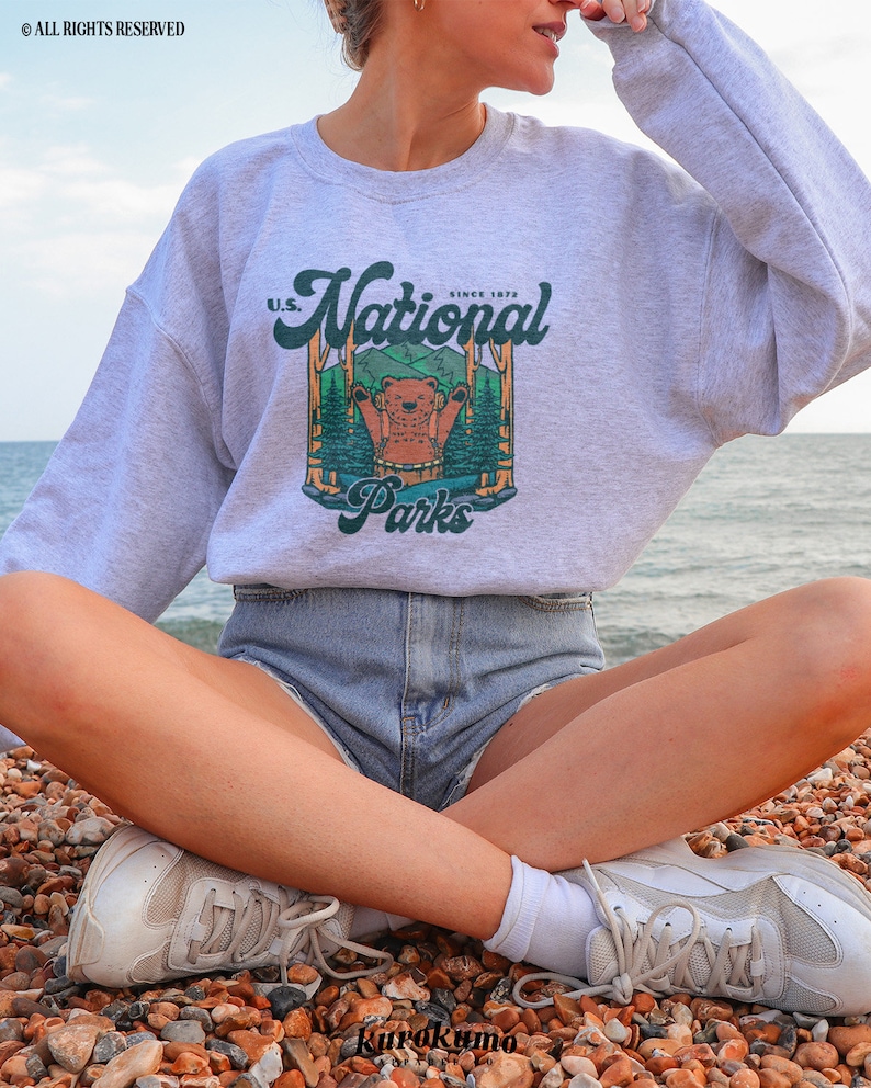 Może przedstawiać: Jasnoszara bluza z nadrukiem przedstawiającym niedźwiedzia w leśnej scenerii oraz napisem "U.S. National Parks Since 1872". Bluza ma okrągły dekolt.