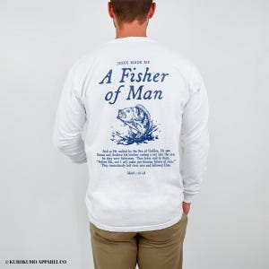 May include: White long-sleeved shirt featuring blue text and a fish illustration. The text says "JESUS MADE ME A Fisher of Man" with a Bible verse underneath. A casual style shirt.