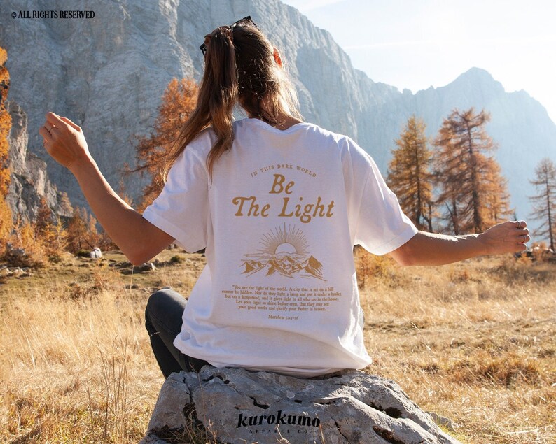 May include: A white t-shirt with a gold graphic design that says "In this dark world, Be the Light". The graphic also includes a sunrise and mountains. The shirt is being worn by a person sitting on a rock in a mountainous landscape.