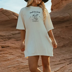Pu&ograve; includere: Una maglietta bianca oversize con una grafica di uno stivale da cowboy che cammina per fede e il testo "walk by faith" e "2 Corinthians 5:7".
