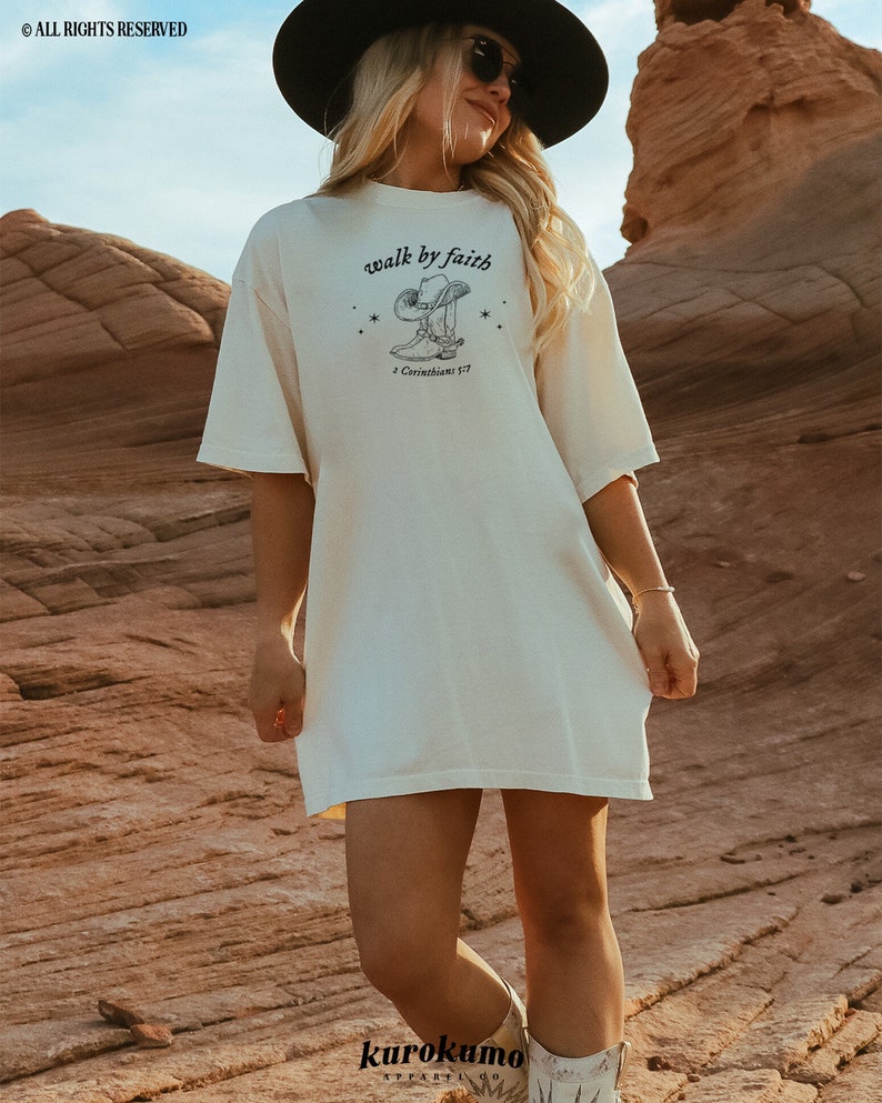 Pu&ograve; includere: Una maglietta bianca oversize con una grafica di uno stivale da cowboy che cammina per fede e il testo "walk by faith" e "2 Corinthians 5:7".
