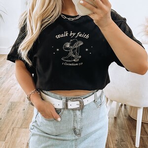 Pu&ograve; includere: Maglietta nera a maniche corte e crop top con una grafica bianca di uno stivale da cowboy con un cappello e il testo "walk by faith" e "2 Corinthians 5:7".