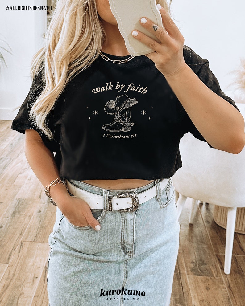 Pu&ograve; includere: Maglietta nera a maniche corte e crop top con una grafica bianca di uno stivale da cowboy con un cappello e il testo "walk by faith" e "2 Corinthians 5:7".