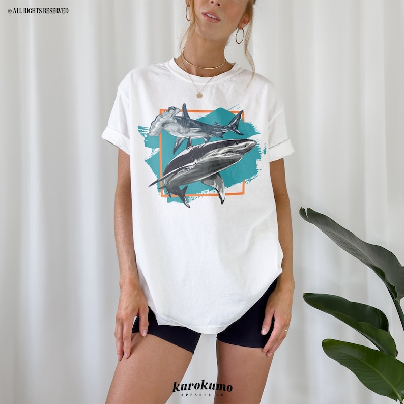 May include: White t-shirt with a graphic of two sharks, one hammerhead and one with a pointed nose, in a turquoise and orange frame. The graphic is on a white t-shirt with a crew neck.