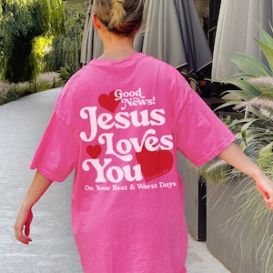 May include: Pink oversized t-shirt with a white graphic that says "Good News! Jesus Loves You" with red hearts and the text "Remember that" in smaller font. The text below the graphic says "On Your Best & Worst Days".