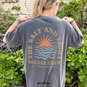 May include: A gray t-shirt with a graphic of a sun setting over the ocean. The text "The Salt and Light Social Club" is printed in a circle around the graphic. The text "Official Matthew 5:13-16 Member" is printed below the graphic.