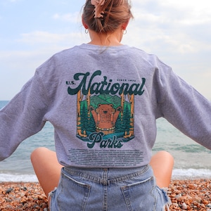 Może przedstawiać: Szara bluza z grafiką niedźwiedzia w leśnej scenerii, z napisem "U.S. National Parks Since 1872". Bluza z długim rękawem ma okrągły dekolt. Osoba siedzi na plaży.