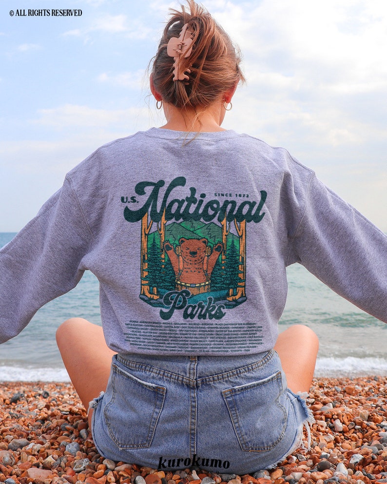 Może przedstawiać: Szara bluza z grafiką niedźwiedzia w leśnej scenerii, z napisem "U.S. National Parks Since 1872". Bluza z długim rękawem ma okrągły dekolt. Osoba siedzi na plaży.