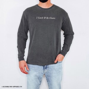 May include: Dark grey long-sleeved shirt featuring the text "I Catch & He Cleans" in white. The shirt has a vintage, washed appearance and is made of a soft material. The model is wearing light blue jeans.