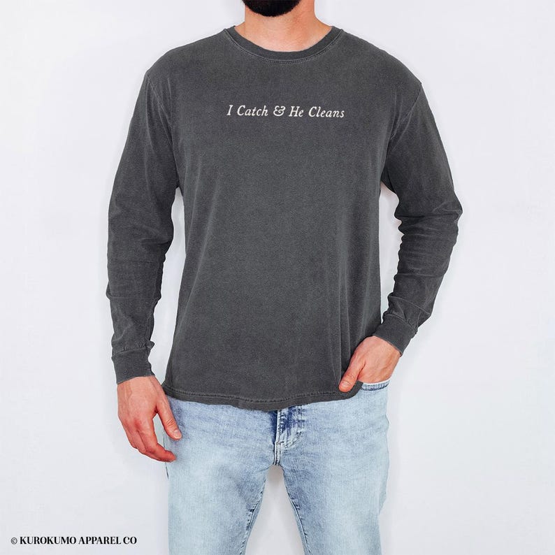 May include: Dark grey long-sleeved shirt featuring the text "I Catch & He Cleans" in white. The shirt has a vintage, washed appearance and is made of a soft material. The model is wearing light blue jeans.
