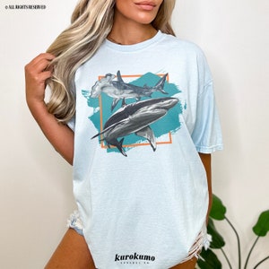 May include: A light blue t-shirt with a graphic print of two sharks, one hammerhead and one great white, in a teal and orange frame. The text "kurokumo apparel co." is printed on the bottom of the shirt.