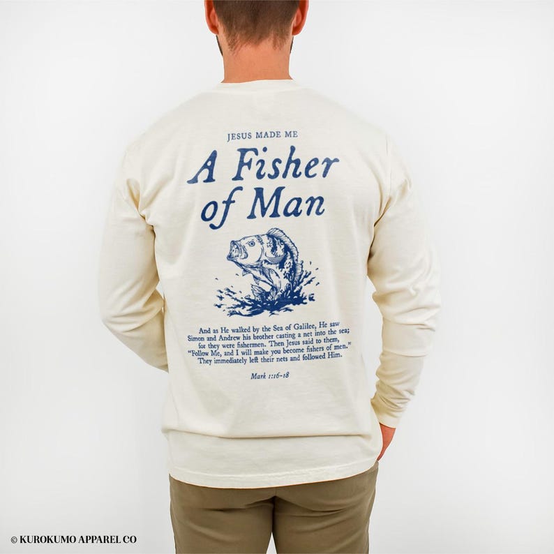 May include: Cream long-sleeved shirt featuring the text "JESUS MADE ME A Fisher of Man" in blue, with a blue fish illustration. A Bible verse is printed below the graphic. The shirt is a neutral colour and suitable for various occasions.