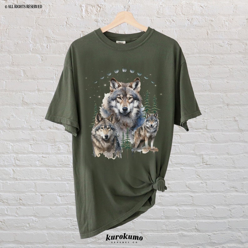 May include: A green t-shirt with a graphic print of three wolves in a forest setting. The wolves are surrounded by trees and a crescent moon. The shirt is made of a soft, comfortable material and is perfect for everyday wear.