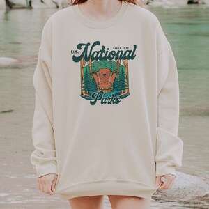 Może przedstawiać: Kremowa bluza z nadrukiem przedstawiającym niedźwiedzia w leśnej scenerii oraz napisem "U.S. National Parks Since 1872." Bluza ma długie rękawy i okrągły dekolt. Nazwa marki "kurokumo apparel co" jest wydrukowana na dole.