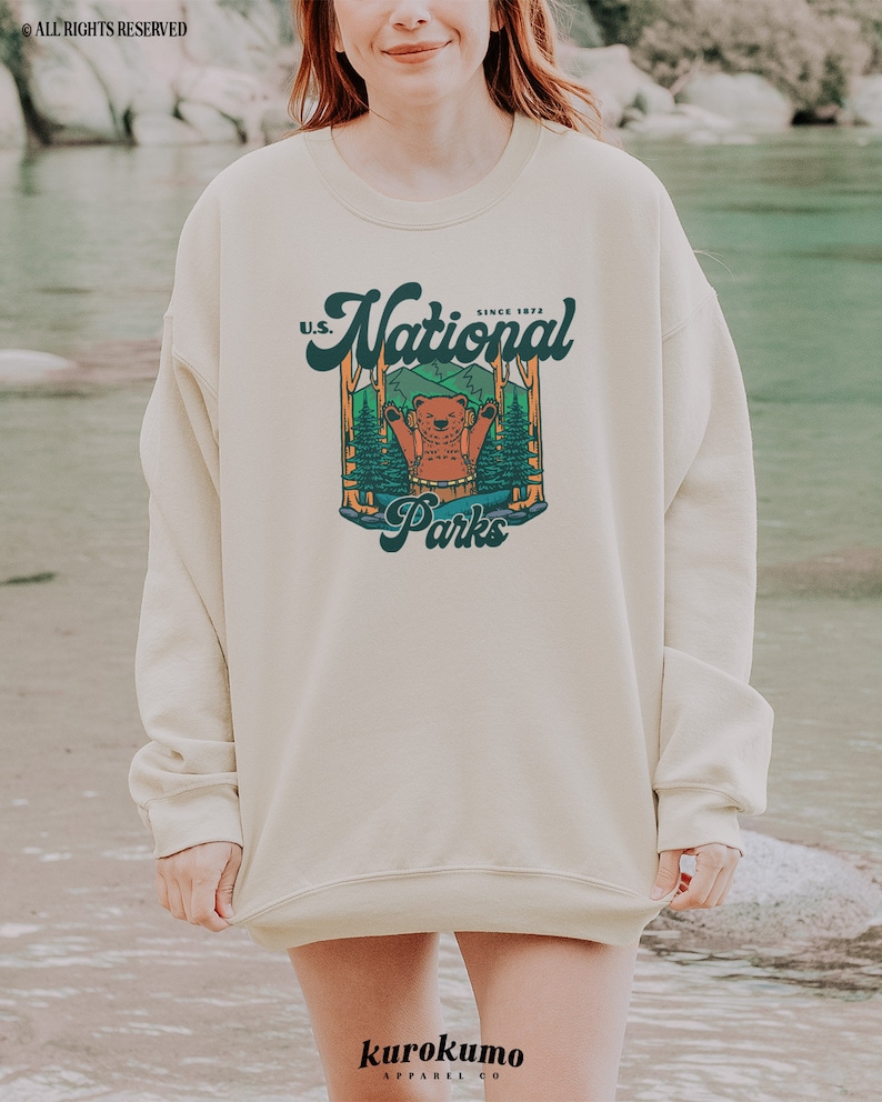 Może przedstawiać: Kremowa bluza z nadrukiem przedstawiającym niedźwiedzia w leśnej scenerii oraz napisem "U.S. National Parks Since 1872." Bluza ma długie rękawy i okrągły dekolt. Nazwa marki "kurokumo apparel co" jest wydrukowana na dole.