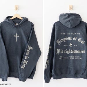 Op de afbeelding: Een donkergrijs hooded sweatshirt met een kruis op de voorkant en de woorden "Jesus is King" op de mouwen. De achterkant van het sweatshirt heeft de tekst "But seek first the Kingdom of God Matt. & 6:31 His righteousness And all these things shall be added to you".