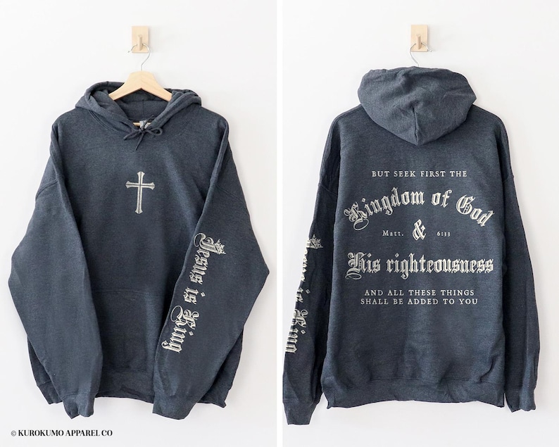 Op de afbeelding: Een donkergrijs hooded sweatshirt met een kruis op de voorkant en de woorden "Jesus is King" op de mouwen. De achterkant van het sweatshirt heeft de tekst "But seek first the Kingdom of God Matt. & 6:31 His righteousness And all these things shall be added to you".