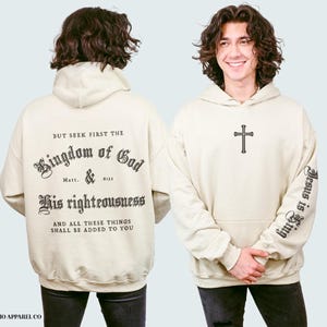 Op de afbeelding: Een beige hoodie met een zwarte print die "But seek first the Kingdom of God Matt. & 6:33 His righteousness And all these things shall be added to you" zegt. De rechtermouw heeft een zwarte print die "Jesus is King" zegt.