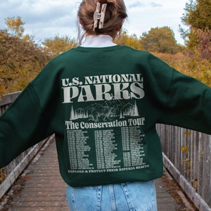 May include: A dark green sweatshirt with the text "U.S. NATIONAL PARKS" and "The Conservation Tour" printed on the back. The design includes a bear graphic and a list of dates and locations. The sweatshirt is worn with blue jeans.