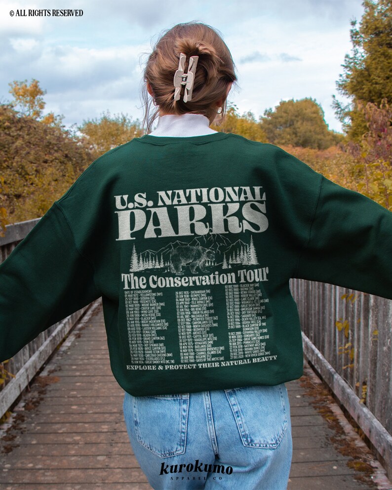 May include: A dark green sweatshirt with the text "U.S. NATIONAL PARKS" and "The Conservation Tour" printed on the back. The design includes a bear graphic and a list of dates and locations. The sweatshirt is worn with blue jeans.
