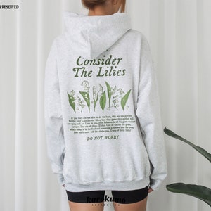 Pu&ograve; includere: Felpa con cappuccio grigia con un disegno floreale verde e il testo "Consider The Lilies" e "Do Not Worry".