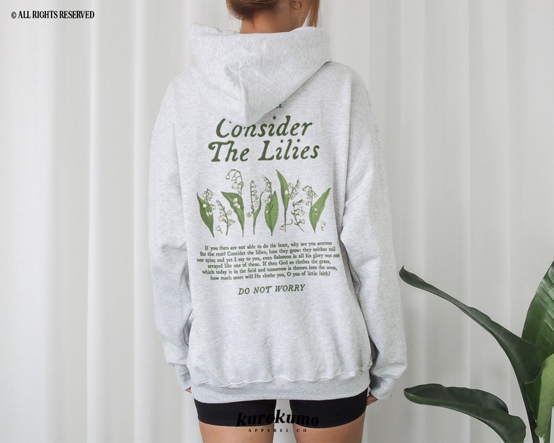 Pu&ograve; includere: Felpa con cappuccio grigia con un disegno floreale verde e il testo "Consider The Lilies" e "Do Not Worry".