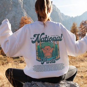 Może przedstawiać: Biała bluza z nadrukiem na plecach przedstawiającym niedźwiedzia w leśnej scenerii, napisem "U.S. National Parks" i listą nazw park&oacute;w. Bluza jest noszona na zewnątrz z g&oacute;rskim tłem.