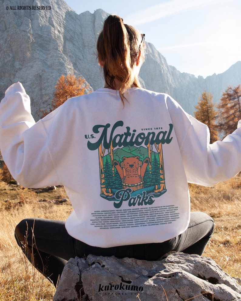 Może przedstawiać: Biała bluza z nadrukiem na plecach przedstawiającym niedźwiedzia w leśnej scenerii, napisem "U.S. National Parks" i listą nazw park&oacute;w. Bluza jest noszona na zewnątrz z g&oacute;rskim tłem.